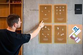 Man interacting with an interactive wall display during the MEA – Opening of MEA at Geneva, pointing at illustrated panels mounted on a concrete wall inside an exhibition space.