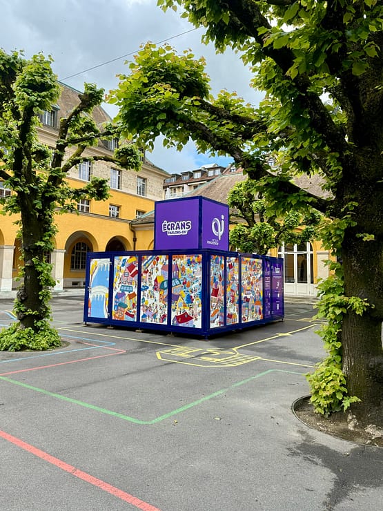 The Action Innocence traveling educational pavilion installed in an outdoor school courtyard, featuring colorful illustrated panels and branded signage, integrated into the environment beneath surrounding trees.