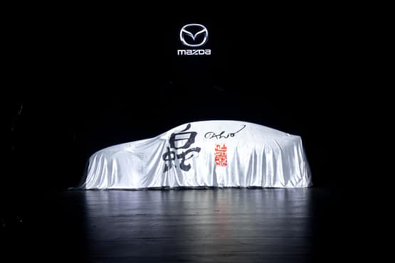 Covered car on stage beneath the Mazda logo during the Mazda – Cinematic reveal of the Mazda 6E.